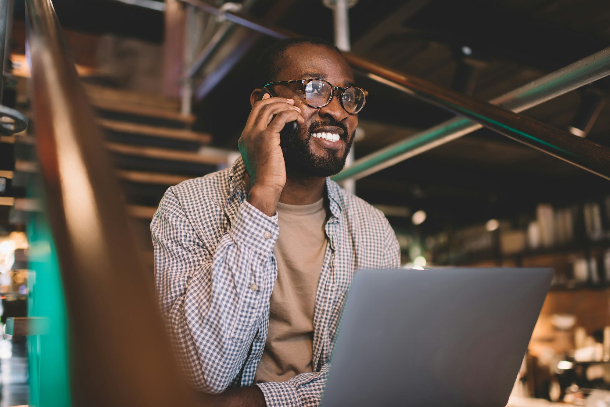 Happy male IT professional in spectacles using laptop and smartphone for call